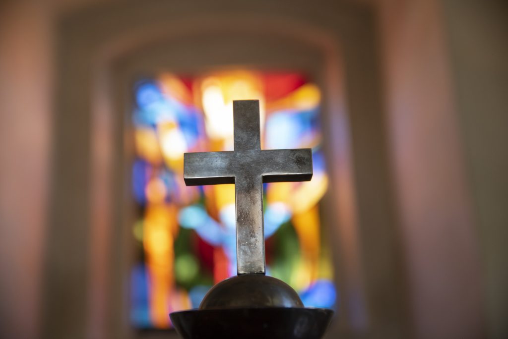 A dim old church interior lit by suns rays penetrating through a colorful stained glass window in the pattern of a crucifix reflecting colors on the floor and a speech pulpit